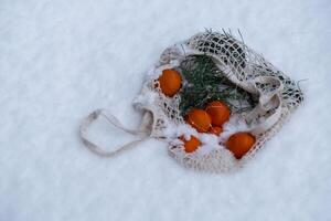 Mesh shopping bag with tangerines in snowy winter outdoor. Go green concept eco-friendly sustainable Christmas. Flat lay in snow background. Responsible celebration photo