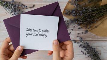 Female hands taking paper card note with text TAKE TIME TO MAKE YOUR SOUL HAPPY from violet envelope. Lavender flower. Top view, flat lay. Concept of mental spiritual health self care wellbeing mindfulness photo