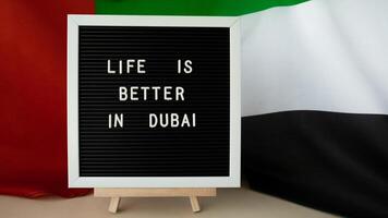 Message LIFE IS BETTER IN DUBAI on background of waving UAE flag made from silk. United Arab Emirates flag with concept of tourism and traveling. Inviting greeting card, advertisement. Dubai welcoming card photo
