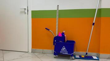 Bucket trolley with detergents and mop for cleaning floor against background of wall and door in corridor, cleanliness in public place, cart on wheels for cleaning service employee, caution wet floor photo