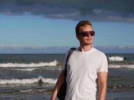 A young man with a backpack, wearing a white T-shirt and sunglasses standing on the seashore at the background of gloomy cloudy sky, with copy space. Tourism, traveling and resort recreation concept photo