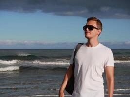 A young man with a backpack, wearing a white T-shirt and sunglasses standing on the seashore at the background of gloomy cloudy sky, with copy space. Tourism, traveling and resort recreation concept photo