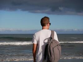 A young man with a backpack, wearing a white T-shirt and sunglasses looking at the sea at the background of gloomy cloudy sky, with copy space. Tourism, traveling and resort recreation concept photo