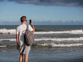 A young man with a backpack, wearing a white T-shirt and sunglasses taking a photo the sea at the background of gloomy cloudy sky, with copy space. Tourism, traveling and resort recreation concept