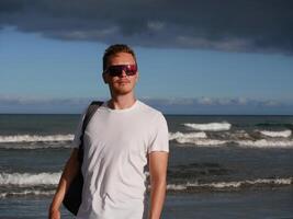 A young man with a backpack, wearing a white T-shirt and sunglasses standing on the seashore at the background of gloomy cloudy sky, with copy space. Tourism, traveling and resort recreation concept photo