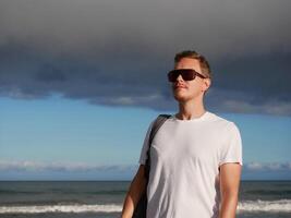 A young man with a backpack, wearing a white T-shirt and sunglasses standing on the seashore at the background of gloomy cloudy sky, with copy space. Tourism, traveling and resort recreation concept photo