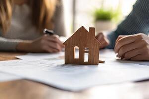 A wooden house model sits on a table with two people working on paperwork in the background. photo
