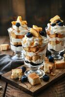 Three dessert parfaits in glass cups with whipped cream, peaches, blueberries, and toasted bread cubes on a wooden serving board. photo