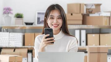 A smiling young woman holding a phone in her hands and looking at the camera. She is sitting at a desk in a home office with cardboard boxes, a laptop, and a plant behind her. photo
