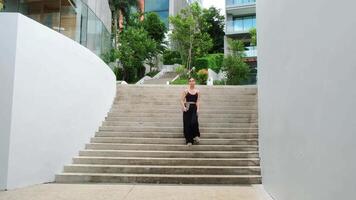 A relaxed woman in black with laptop walking by a modern office, enjoying the green city atmosphere. The perfect moment to blend work and everyday life in a comfortable setting video