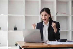 Enthusiastic Business Woman Working on Laptop in Modern Office, Celebrating Success, Professional Female in Formal Attire, Productive Work Environment, Corporate Achievement, Office Desk Setup photo