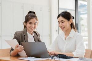 Young Businesswoman and Colleague Collaborating on Project in Modern Office Setting with Documents and Laptop, Smiling and Engaged in Productive Work Environment photo