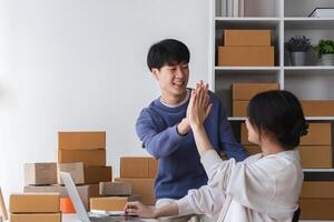 Happy Couple Running a Small Online Business from Home, Surrounded by Cardboard Boxes and Working on a Laptop, Celebrating Success with a High Five in a Bright Modern Workspace photo