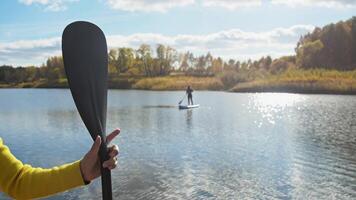 In distance, young woman stands on surfboard in middle of lake or river. In foreground on left, woman's hand, dressed in yellow sweater, holds rowing paddle. pond is surrounded by trees, leaves of which acquire bright autumn shades. video
