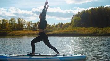 Young, fit woman practices yoga while standing on surfboard, which is positioned on water's surface. She executes movements with precision, maintaining her balance effortlessly. video