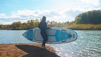 On serene autumnal day, young woman takes to water, launching her stand-up paddleboard in preparation for embarking on her practice of yoga on surface of water. video