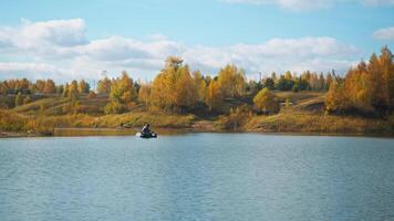 A lone fisherman in small inflatable boat floats in the center of lake, surrounded by an autumnal landscape with backdrop of golden autumn forest. fisherman is engaged in fishing. video
