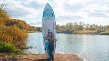 Young, lovely woman stands in front of large, floating surfboard against backdrop of tranquil pond on crisp autumn day. video