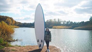 Woman of Asian descent, enjoys activity of surfing on stand-up paddleboard, stands next to board, preparing to depart for water. Woman's smile and gaze into camera lens convey sense of joy and confidence. video