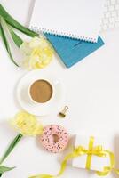 Notebook for daily planning with breakfast serving on white background, cup of coffee, donut, flowers and gift box with bow. Concept of daily planning of life, work, study. Mother's Day. Copy space photo
