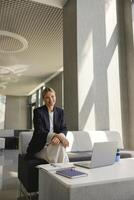 Smiling business woman working on laptop sitting on background of modern office and looks camera photo