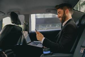 Smiling entrepreneur working on laptop and use mobile phone while going to airport by car photo