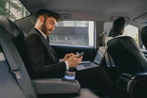 Busy male analyst working on laptop and use mobile phone while going to airport by car photo