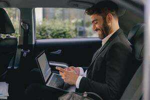 Smiling entrepreneur working on laptop and use mobile phone while going to airport by car photo