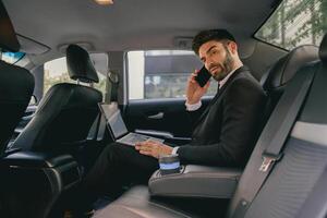 Handsome businessman in suit using personal computer and making phone call in taxi photo