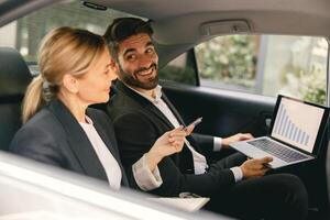 Business colleagues working on laptop while sitting in car backseats on way to office photo
