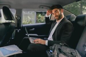 Handsome businessman in suit working on laptop sitting on car backseats and looks camera photo