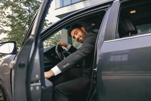 Smiling businessman in suit looking at camera while sitting in driver seat with opened door photo