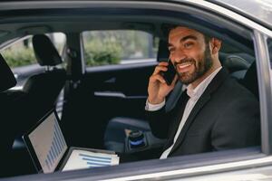 Handsome smiling businessman in suit using personal computer and making phone call in taxi photo