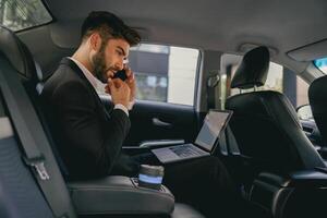Busy entrepreneur working on laptop and talking on phone while going to airport by car photo
