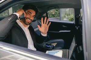 Smiling businessman in suit with laptop making phone call sit in taxi and waving Hi in open window photo