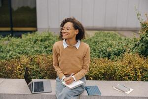 Smiling business woman working laptop and making notes sitting on background of office building photo