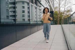 Smiling business woman is talking phone while standing with laptop on modern building background photo