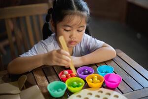 Asian young girl is sitting at a table with a variety of colorful plastic cups. She is using a spoon to scoop out the contents of the cups and place them into a bowl photo