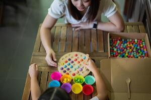Asian woman and a child are sitting at a wooden table, playing a game with colorful balls. The woman is helping the child with the game, and they are both smiling and enjoying themselves photo