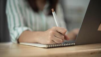 Asian woman is writing in a notebook with a pen. She is sitting at a desk with a laptop in front of her. The scene suggests that she is working or studying, possibly taking notes or writing a report photo