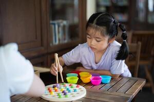 Asian young girl is playing with a game that has a lot of colorful balls. She is using chopsticks to pick up the balls. A woman is watching her photo