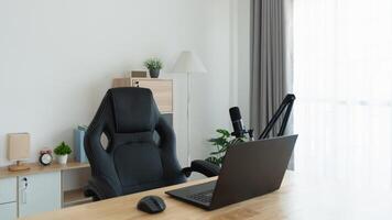 A black office chair sits in front of a laptop computer. The chair is positioned in front of a window, and a potted plant is nearby. The room appears to be a home office, with a clock on the wall photo