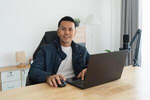 Asian man is sitting at a desk with a laptop and a mouse. He is smiling and he is enjoying himself photo