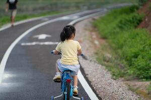 Asian young girl is riding a blue bicycle down a road. The road is lined with grass and there is a man walking behind her. The scene is peaceful and calm, with the girl enjoying her ride photo