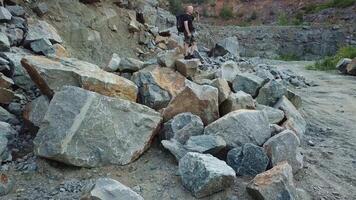 A tourist in shorts and a backpack is standing on a huge stone in the middle of a quarry and looking into the distance to the surrounding landscape. Camera motion up. Aerial view. video