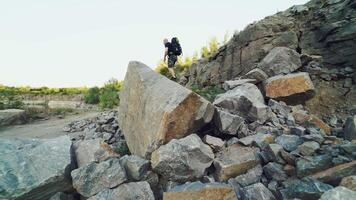 A traveler in shorts and with a backpack climbs onto a huge stone and sits on it amid a quarry with a lake in the center. video