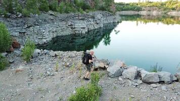 A traveler in a black T-shirt with a backpack on his shoulders walks along the waterside of the lake near the quarry with stones and trees. Wonderful summer landscape. video