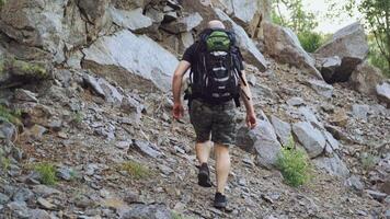 A traveler with a backpack on his shoulders rises to the top of the mountain with stones in the afternoon in warm weather. Tourism. video