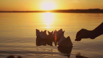 Female's hand putting one paper boat on the water and it joins to other ones at sunset. New origami ship adding to the rest during beautiful sunset with the sun reflection in the lake video