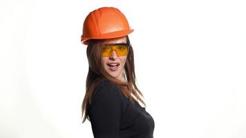 A beauty with long brown hair in an orange helmet and glasses is holding a hammer in her hand and posing with it on a white background in the studio. video